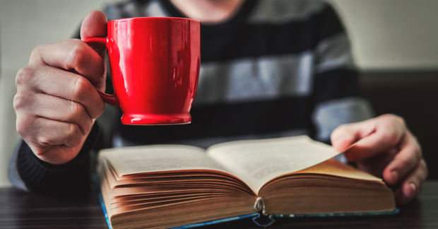 When Person holding a bright red mug while reading an open book, with the mug in sharp focus and the reader blurred in the background.