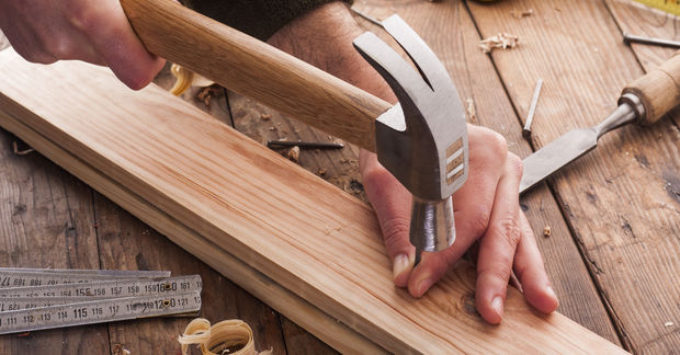 Put Hands using a hammer to drive a nail into a wooden board surrounded by woodworking tools and measuring tape on a wooden work surface.