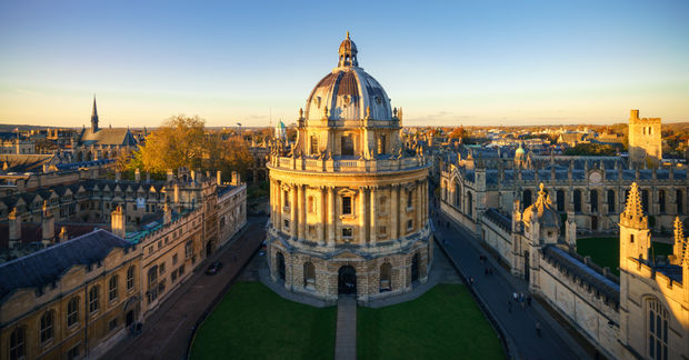 An aerial view of the University of Oxford, Aerial view of a grand domed classical stone building illuminated by golden hour sunlight, surrounded by historic architecture and green courtyard, with church spires visible in background.