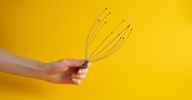 A hand holds a wire scalp massager with metal prongs tipped in small balls against a bright yellow background.