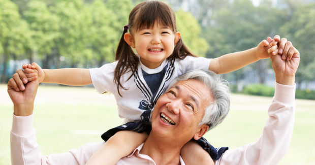 Grandfather and granddaughter laughing together outdoors, with child sitting on his shoulders with arms spread wide in a park setting.