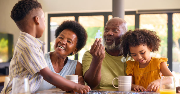 Multi-generational family laughing together around a table playing a board game with mugs and natural light from windows in the background.